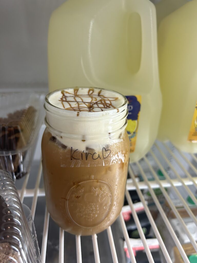 This is a photo of a mason jar full of light brown iced latte with cream coloured cold foam and a light brown caramel drizzle on top. The mason jar is in a fridge.
The iced latte was for my coworker so the mason jar has a piece of clear tape with her name "Kira" and a love heart written on it.
Photo credits: Me (Kennedy Mosses)