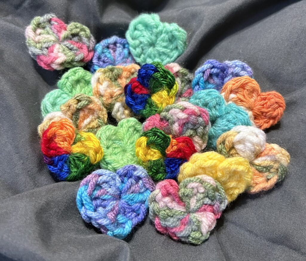 A pile of roughly 20 multi-coloured mini crocheted hearts laying on a gray sheet backround